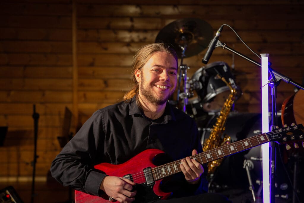 Smilende gitarist spiller rød elgitar på scenen, med trommesett, saksofon og mikrofon i bakgrunnen.