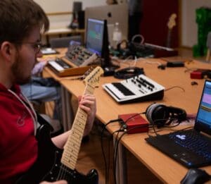 Ung mann spiller elektrisk gitar ved hjemmestudio med laptop, lydgrensesnitt, hodetelefoner og MIDI-synth på bordet.