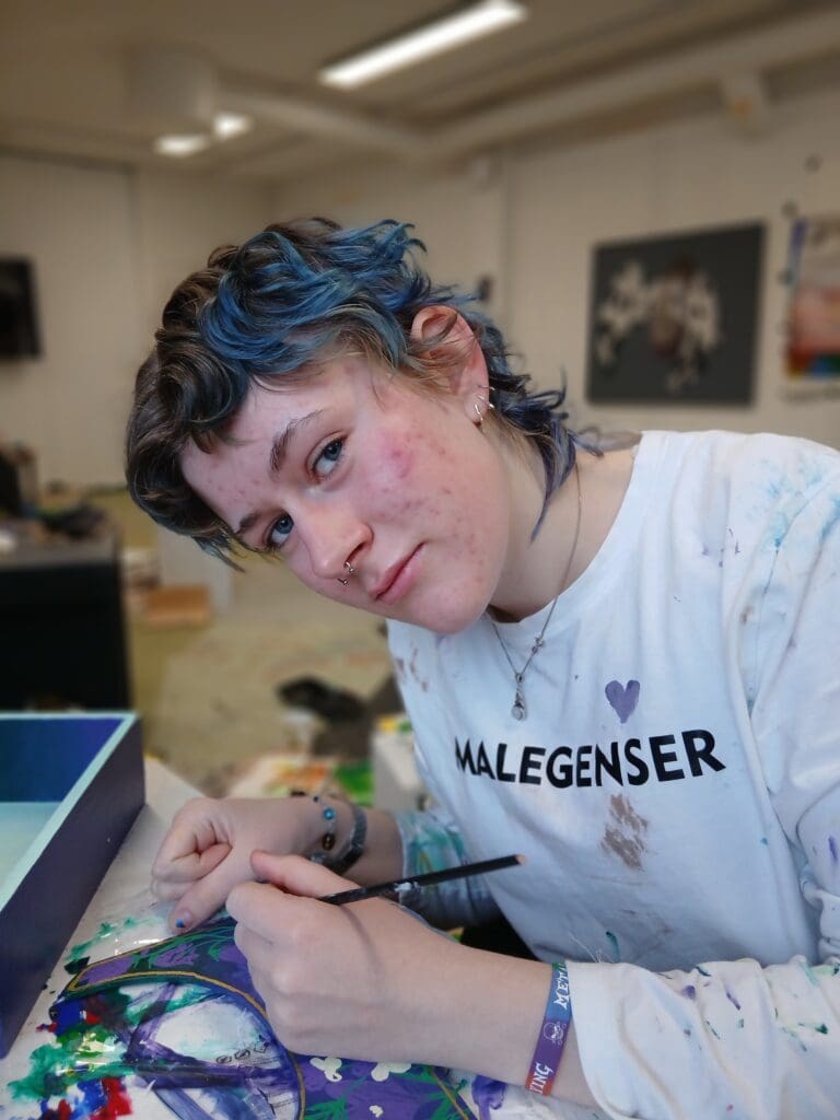 Ung kunstner med blått hår maler i atelier, holder pensel og har t-skjorte med teksten MALEGENSER.