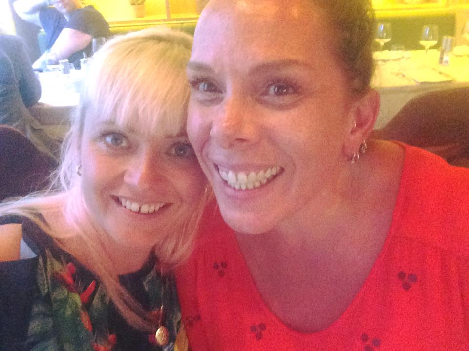 To kvinner tar en glad selfie ved middagsbord på restaurant, smilende i varm belysning.