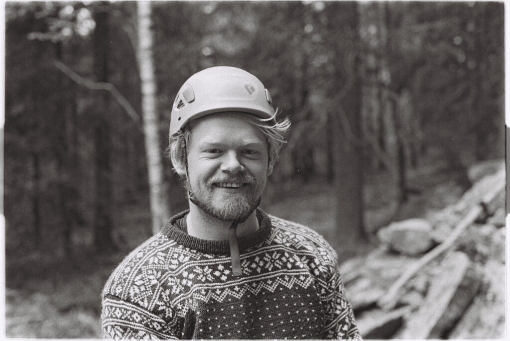 Smilende person i klatrehjelm og mønstret ullgenser i skog, sorthvitt utendørsportrett.