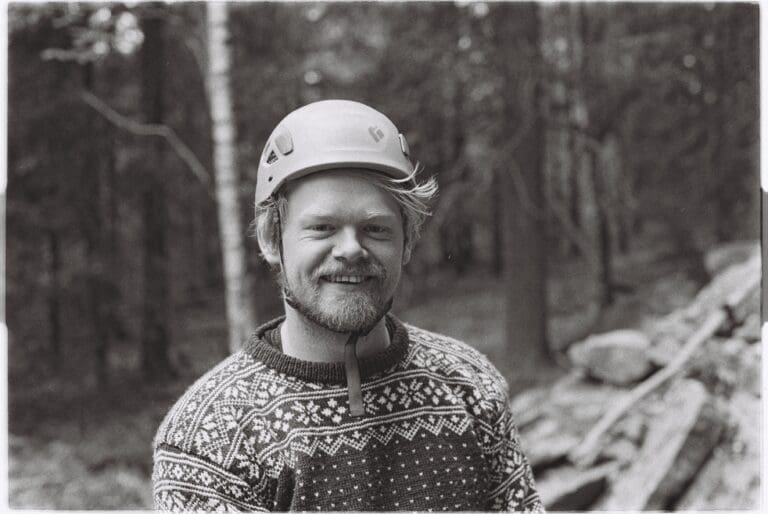 Smilende person i klatrehjelm og mønstret ullgenser i skog, sorthvitt utendørsportrett.