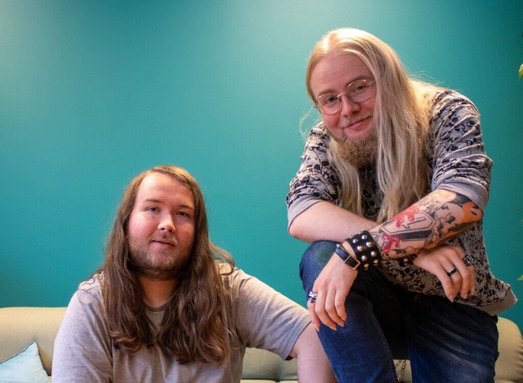 To personer med langt hår sitter på sofa foran turkis vegg; en med tatoveringer og briller smiler mot kamera.