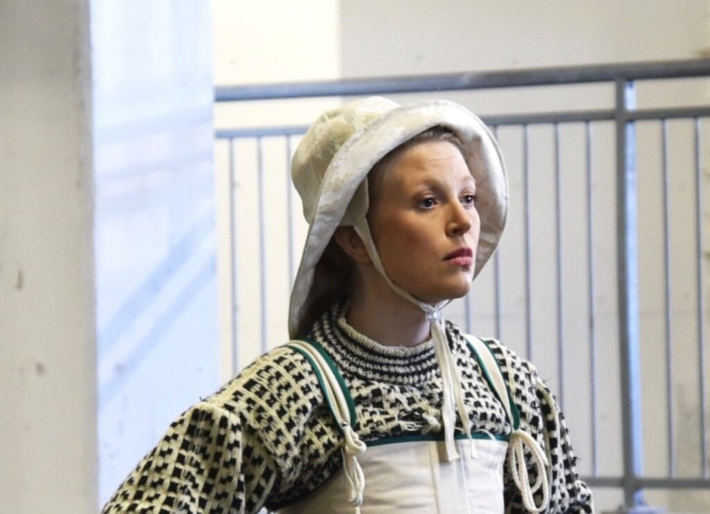 Kvinne i tradisjonell norsk drakt med hvit kyse og mønstret strikkegenser, poserer innendørs ved metallrekkverk.